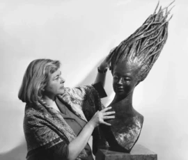 L'artista con una sua scultura Fotografia in bianco e nero, Lalla Vanzella in piedi, di fronte ad una sua scultura in bronzo dai lunghissimi capelli.